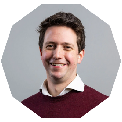 Smiling man with dark hair wearing a maroon sweater and white shirt, posing against a gray background.
