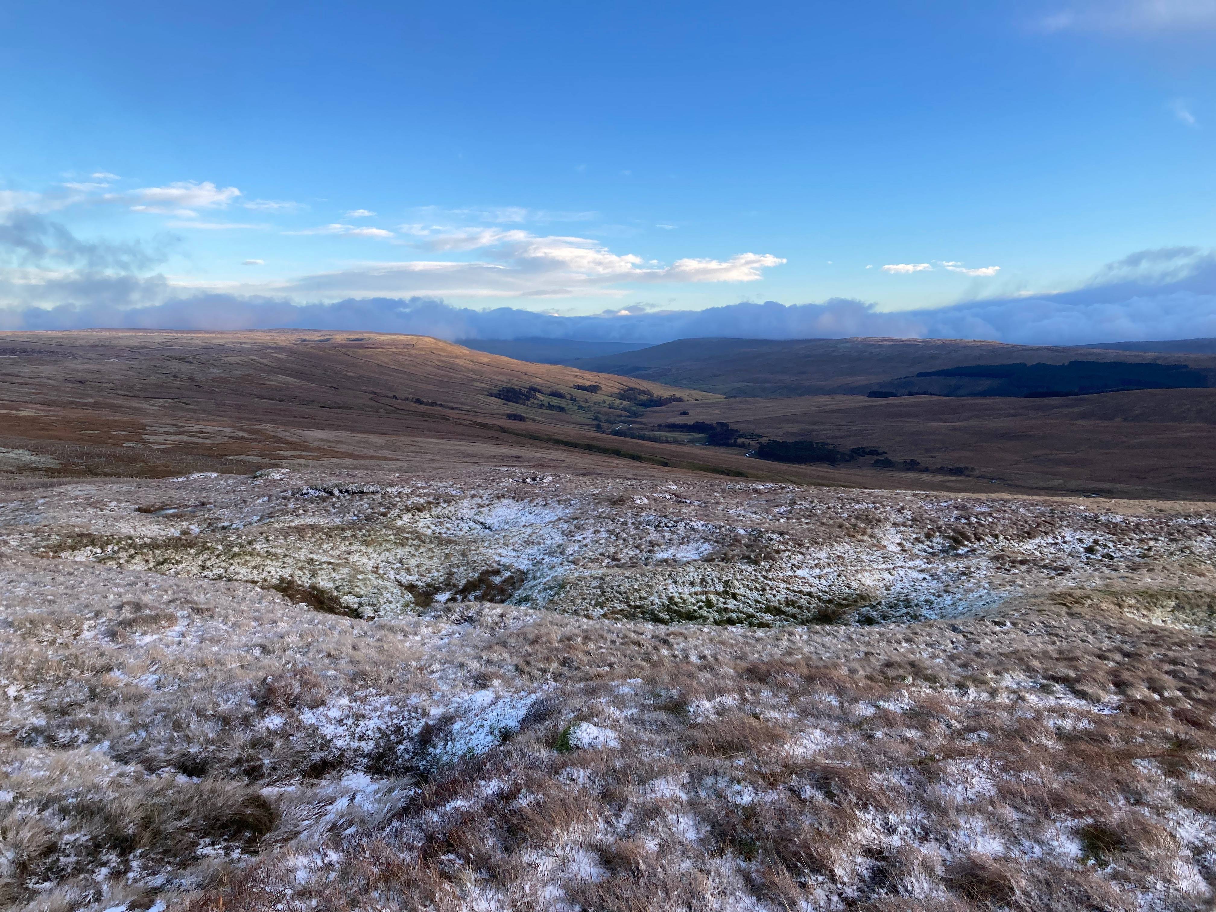 A vast, snow-dusted moorland under a clear blue sky, with rolling hills and distant mountains on the horizon.