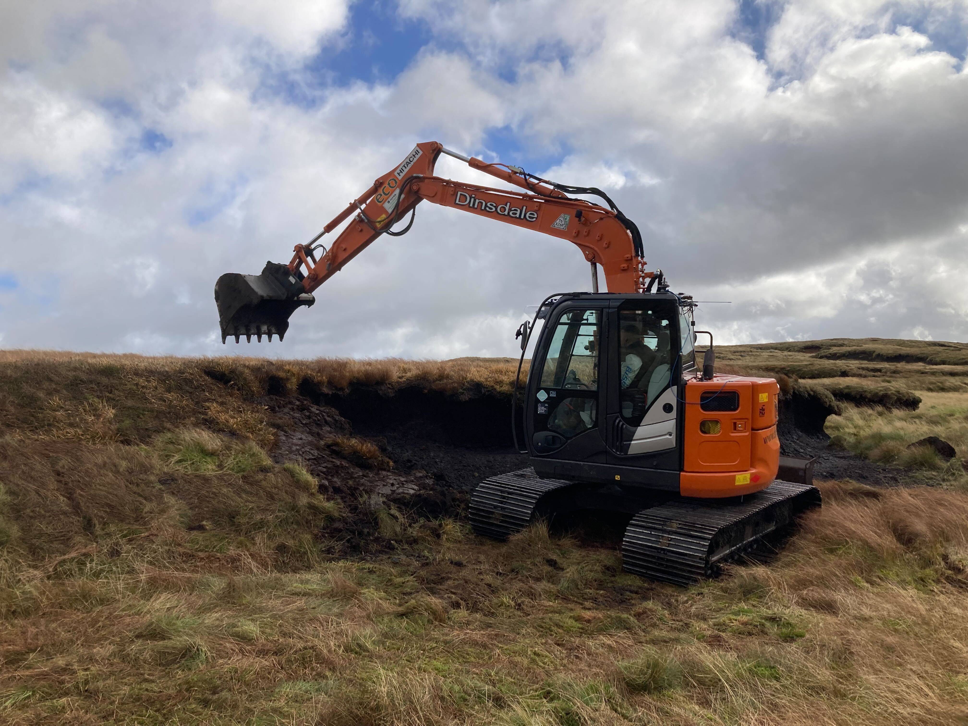 An orange excavator digging into a grassy, hilly landscape under a cloudy sky.