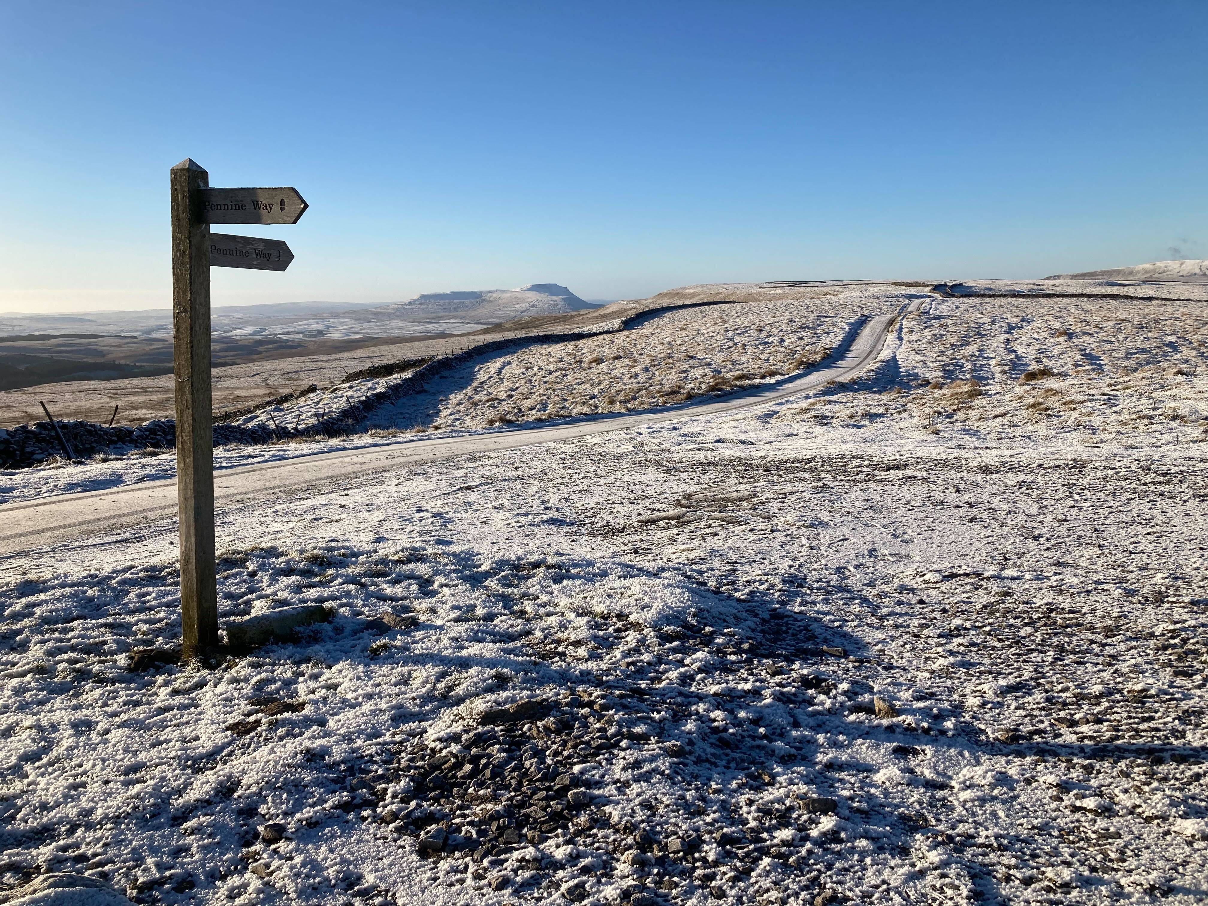 Snow-covered landscape with a wooden signpost pointing in two directions, under a clear blue sky. A winding path stretches into the distance.