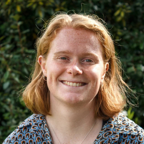 Smiling person with short red hair, wearing a patterned shirt, stands in front of green foliage.