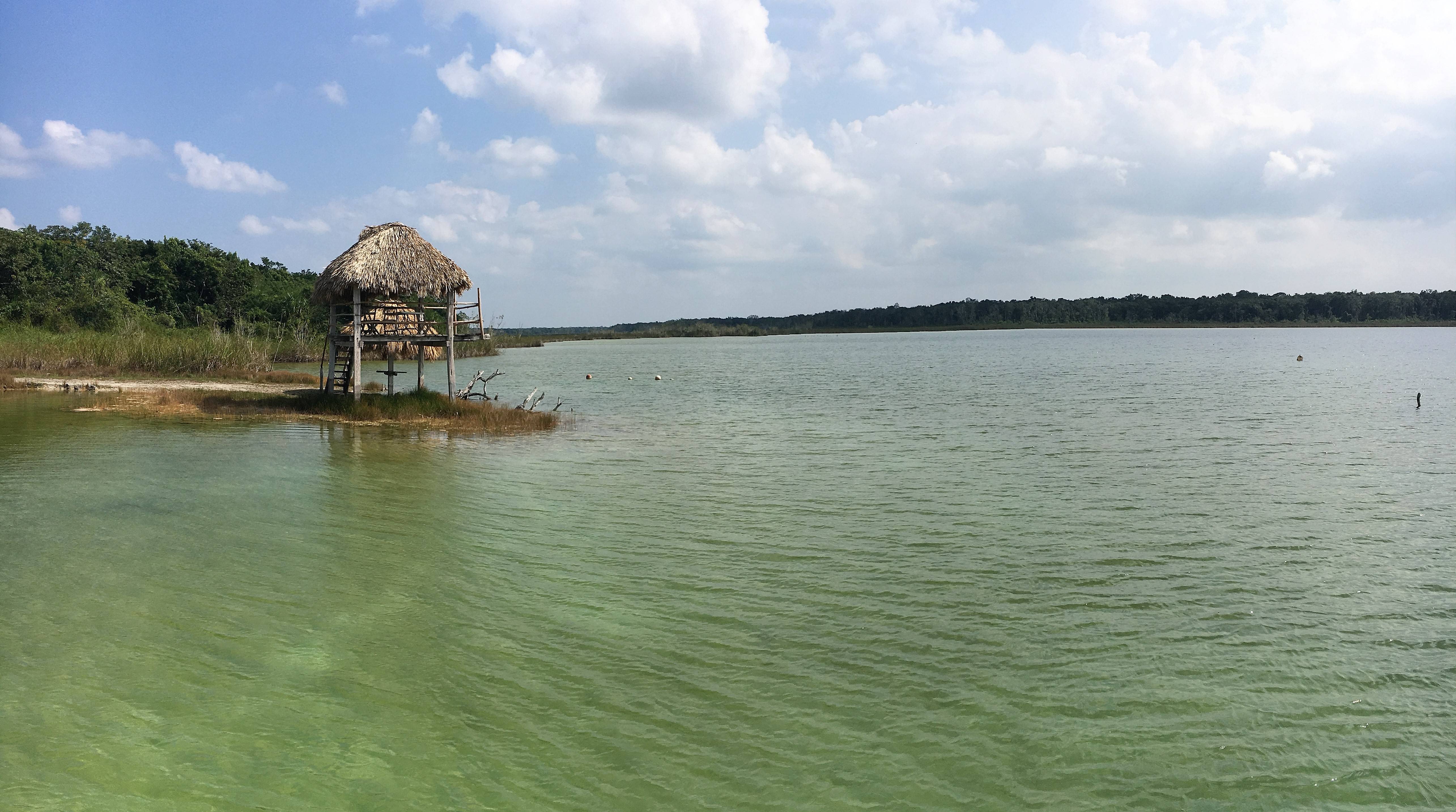 A small thatched hut on stilts stands by the edge of a calm, green lake, with a forested shoreline under a partly cloudy sky.