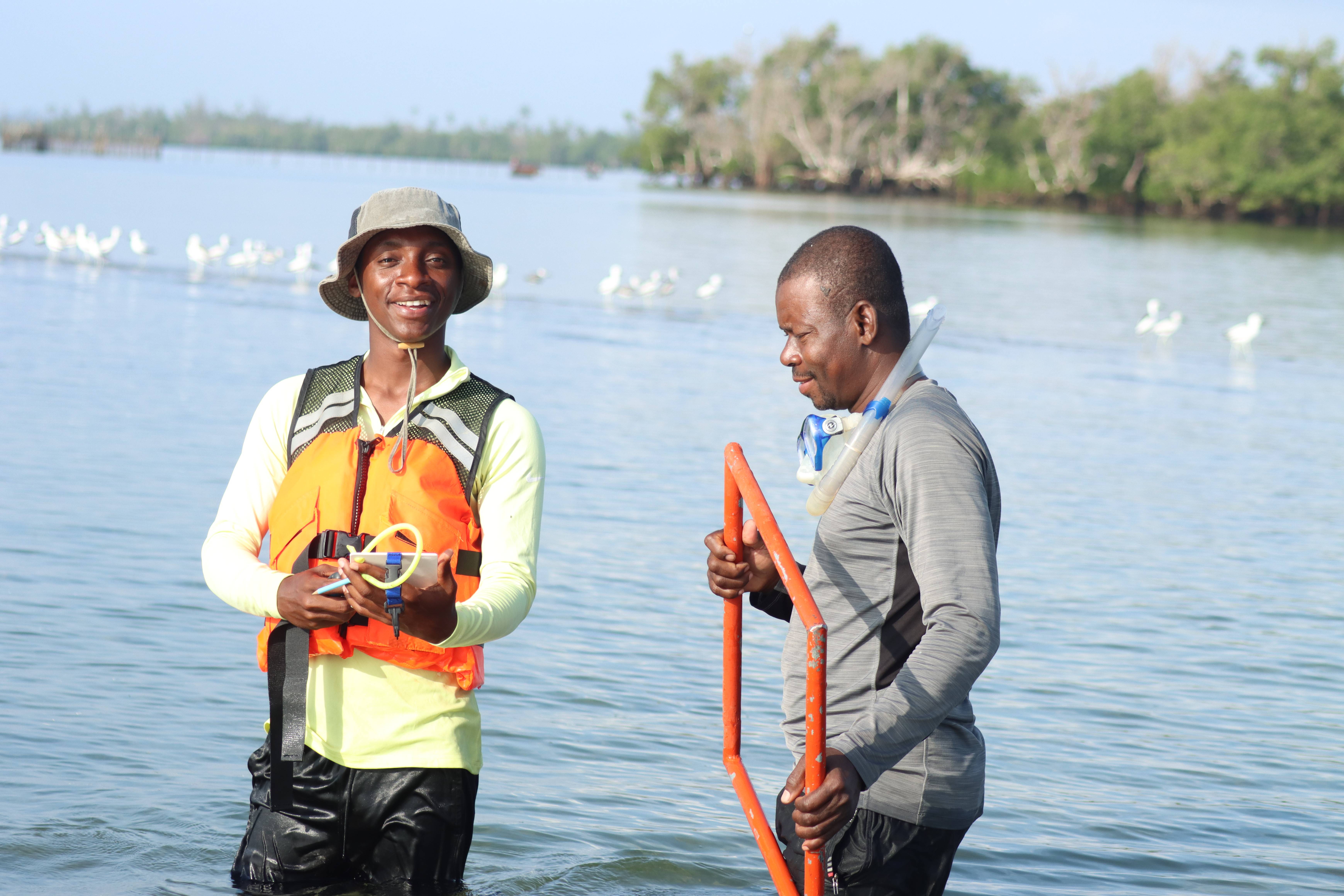 Two men in water, one in an orange vest holding a device, the other with a snorkel, surrounded by seagulls and trees.
