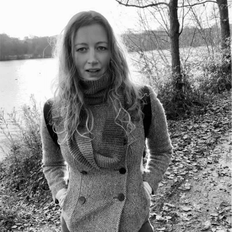 Woman with long hair and a scarf stands on a leaf-covered path by a lake, trees in the background; black and white image.