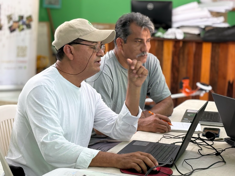 Two men sitting at a table, focused on laptops. One gestures while speaking. Papers and a green wall are in the background.