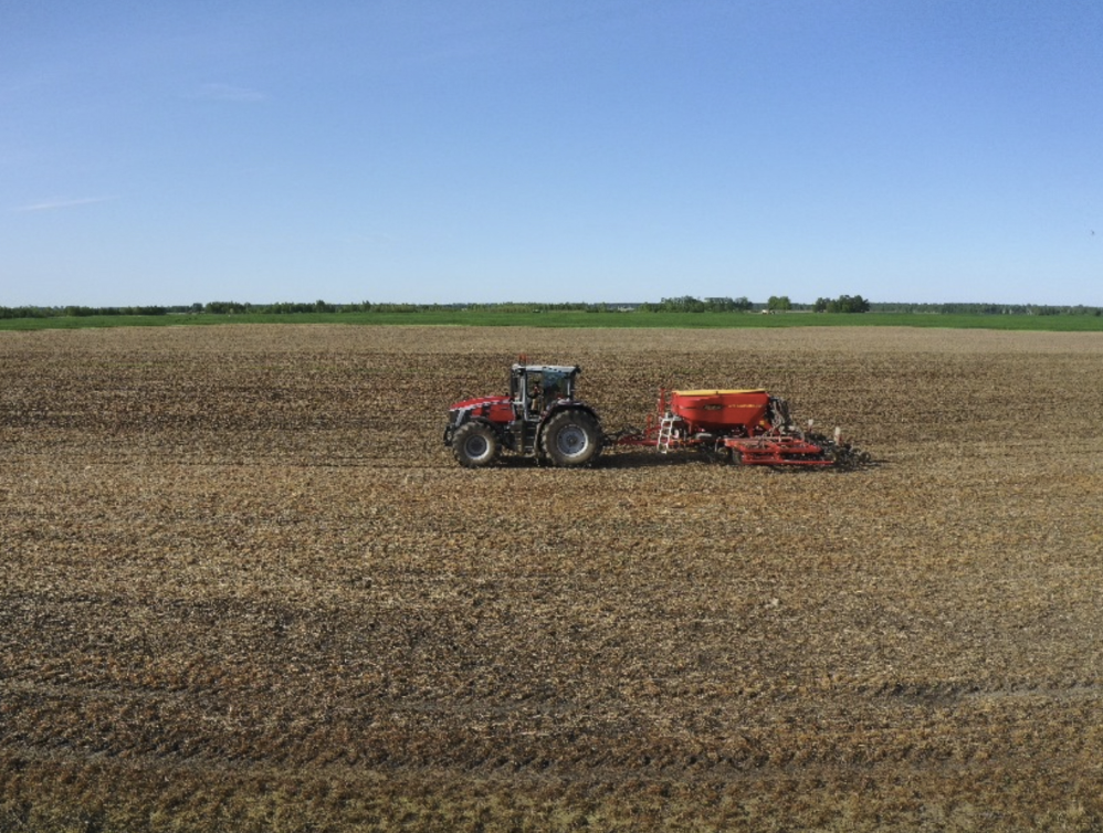 A red tractor pulling a seeder, working on a vast, brown field under a clear blue sky.