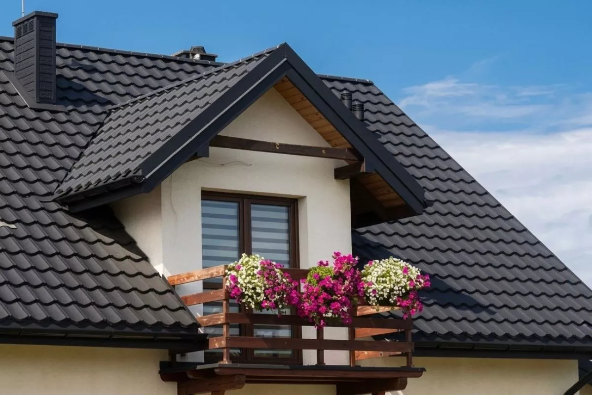 A small balcony with colorful flowers is attached to a cream-colored house with a dark tiled roof under a clear blue sky.