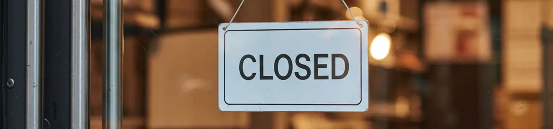 A "Closed" sign hangs on a glass door of a dimly lit store, with shelves and boxes visible in the background.