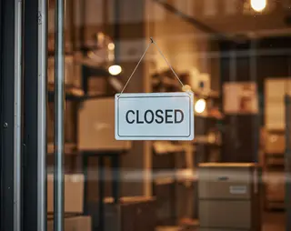 A "Closed" sign hangs on a glass door of a dimly lit store, with shelves and boxes visible in the background.