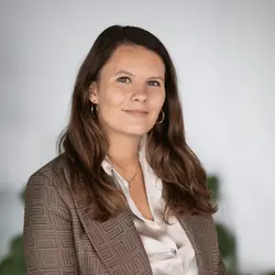 Vrouw met lang bruin haar in een bruine blazer en witte blouse staat voor een witte achtergrond met groene planten.