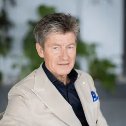 Man met grijzend haar draagt een beige colbert en donker overhemd, zittend in een lichte ruimte met groene planten op de achtergrond.