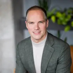 Man met kort haar en grijze blazer zit op een stoel in een moderne omgeving met planten op de achtergrond.