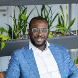Man met bril en blauwe blazer glimlacht, zittend voor planten in een moderne ruimte.