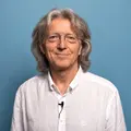 Man met halflang grijs haar en bril draagt een witte blouse en kijkt glimlachend naar de camera, tegen een blauwe achtergrond.