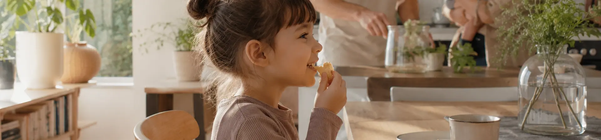 A family in a bright kitchen; a girl eats at the table; a couple in the background interacts with a baby, surrounded by plants and decor.