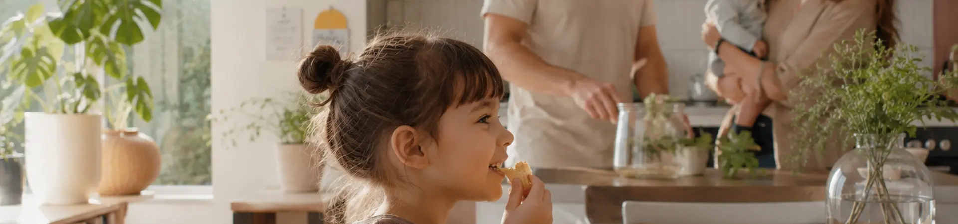 A family in a bright kitchen; a girl eats at the table; a couple in the background interacts with a baby, surrounded by plants and decor.