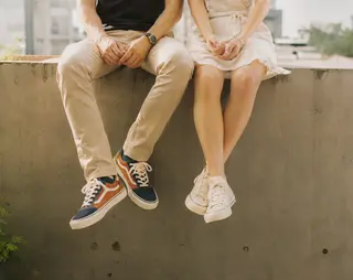 Two people sitting on a ledge, legs dangling. One wears colorful sneakers and beige pants, the other is in a white dress and white sneakers.