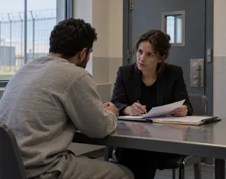 Een vrouw in een zwarte blazer zit tegenover een man in een grijze trui aan een tafel, met documenten in een verhoorkamer.