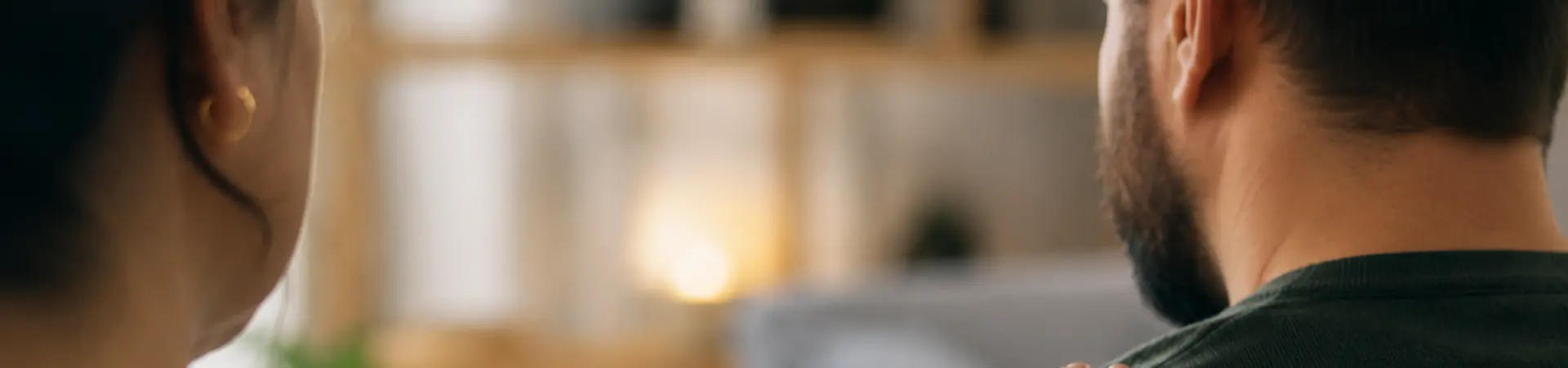 A person places a comforting hand on another's shoulder, both seated in a cozy room with blurred shelves and soft lighting in the background.
