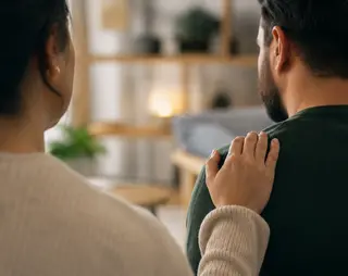 A person places a comforting hand on another's shoulder, both seated in a cozy room with blurred shelves and soft lighting in the background.
