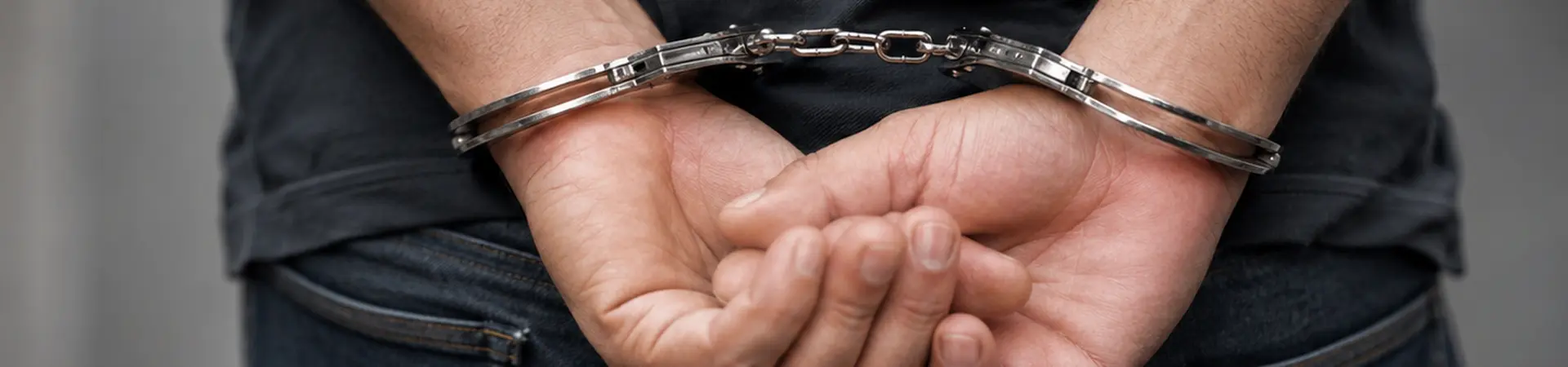 Person in jeans and a dark shirt with hands cuffed behind their back, standing against a blurred background.