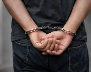 Person in jeans and a dark shirt with hands cuffed behind their back, standing against a blurred background.