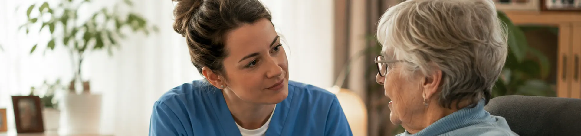 A caregiver in blue scrubs sits beside an elderly woman in a cozy room, engaging in a warm conversation. A walker sits next to them.