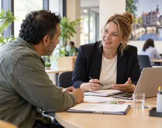 Een vrouw en man in gesprek aan een tafel met documenten en een laptop, in een kantooromgeving met groene planten.