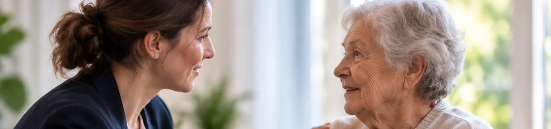 Een jonge vrouw praat vriendelijk met een oudere vrouw in een rolstoel, terwijl ze haar schouder aanraakt. Ze glimlachen naar elkaar.