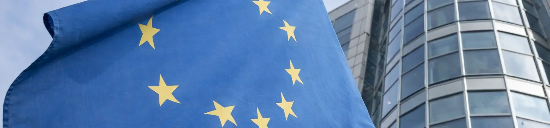 European Union flag with yellow stars on a blue field, waving in front of a modern glass office building against a cloudy sky.