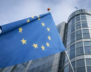 European Union flag with yellow stars on a blue field, waving in front of a modern glass office building against a cloudy sky.