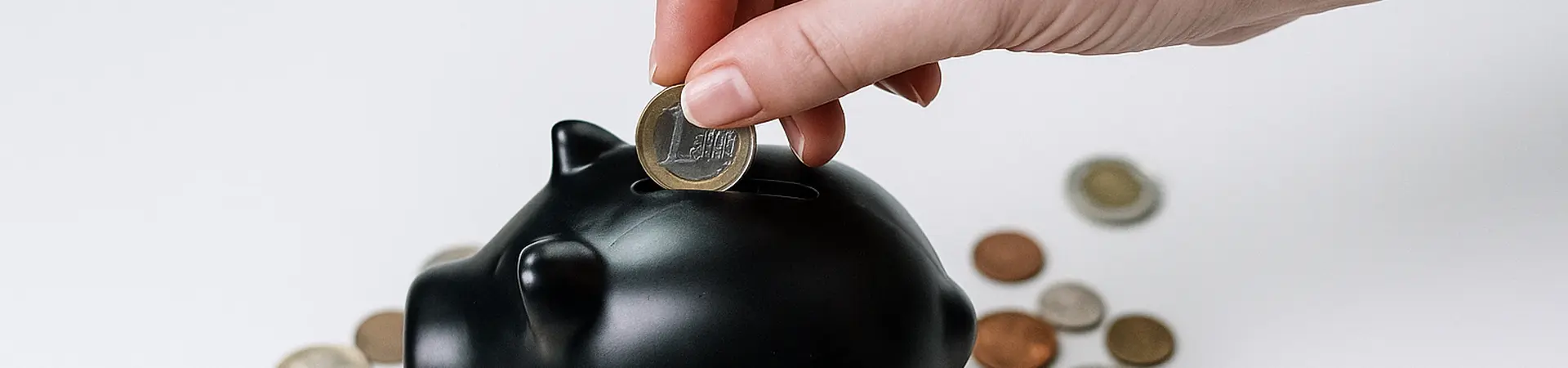 Hand placing a coin into a black piggy bank surrounded by scattered coins on a white surface.