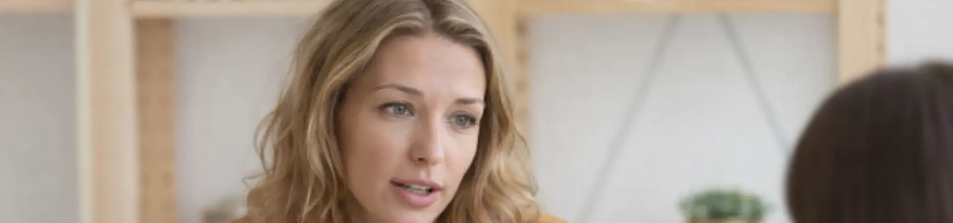 Woman in a mustard blouse gesturing while conversing with another person at a table, shelves with books and plants in the background.