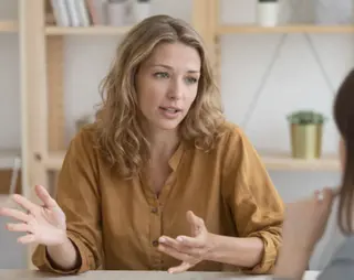Woman in a mustard blouse gesturing while conversing with another person at a table, shelves with books and plants in the background.