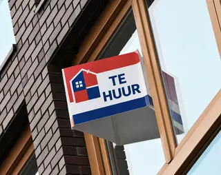 A rectangular sign with a house icon and "Te Huur" text, hangs on a building's exterior. The sign is red, white, and blue.