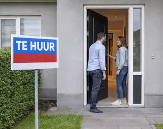 A man and woman stand at the entrance of a house with a "Te Huur" sign, indicating it's for rent. The woman holds the door open.