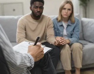 A couple sits holding hands on a sofa, attentively listening to a person with a clipboard and pen, suggesting a counseling session.