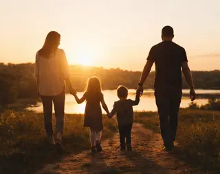 A family of four holding hands, walking towards a sunset by a lake, with trees in the background.