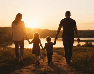 A family of four holding hands, walking towards a sunset by a lake, with trees in the background.