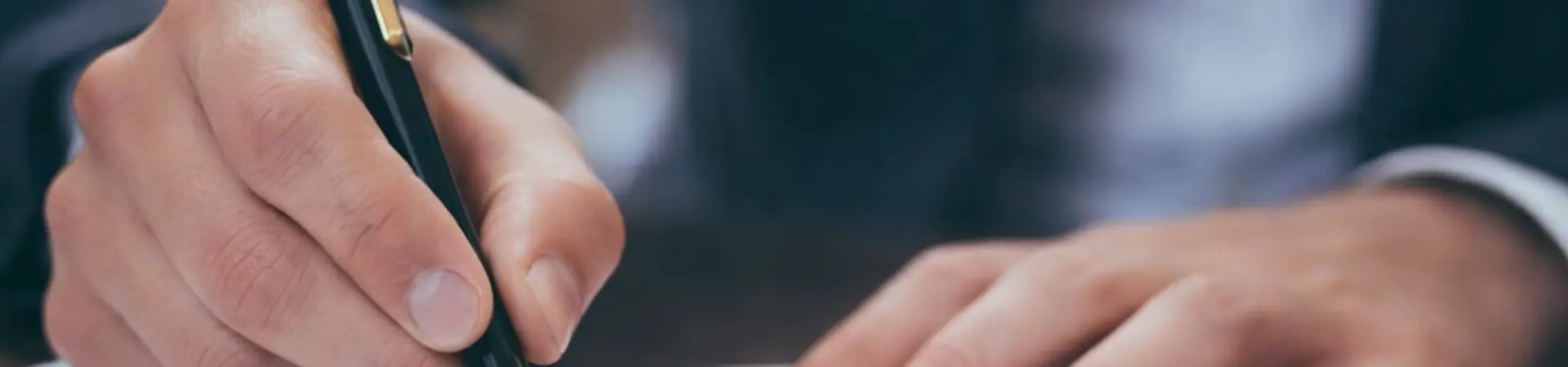A person in a suit signing a document with a black and gold pen on a wooden desk.