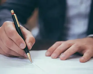 A person in a suit signing a document with a black and gold pen on a wooden desk.
