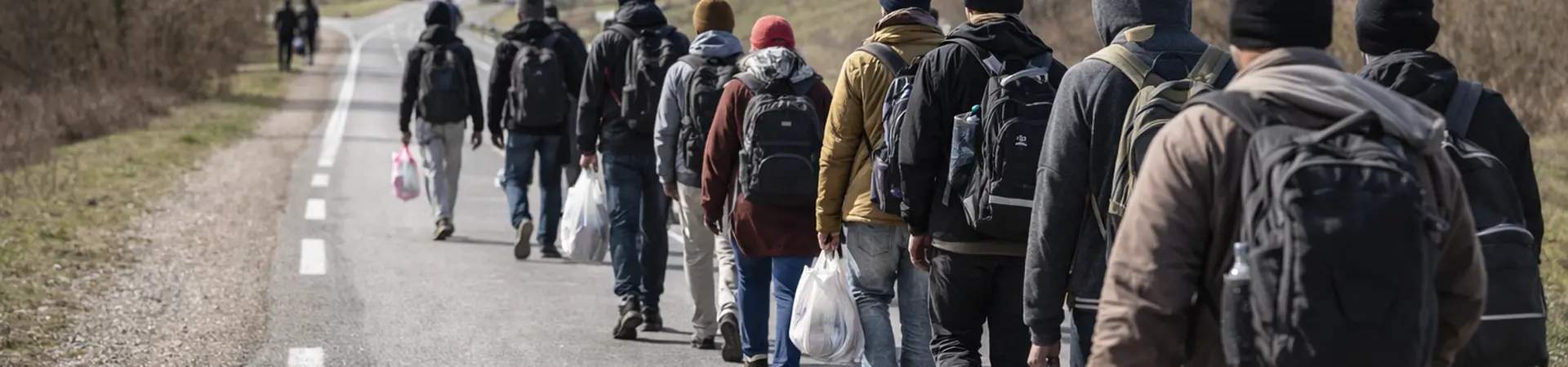 A group of people with backpacks and bags walk along a rural road, surrounded by fields and bare trees under a cloudy sky.