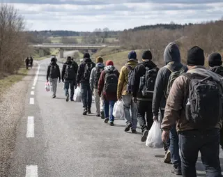 A group of people with backpacks and bags walk along a rural road, surrounded by fields and bare trees under a cloudy sky.