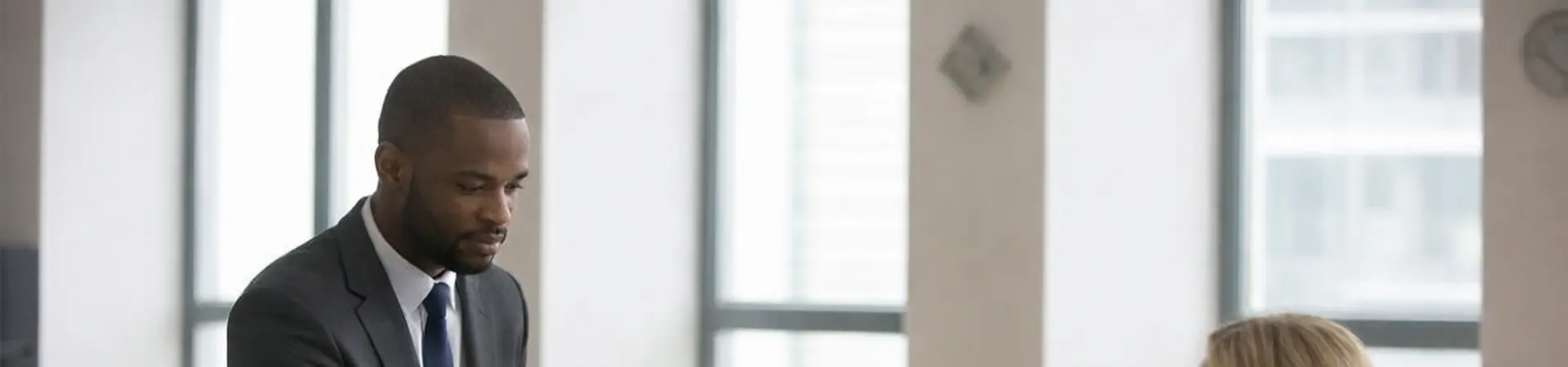 A man in a suit hands a document to a woman seated at a desk in an office with large windows.