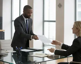 A man in a suit hands a document to a woman seated at a desk in an office with large windows.