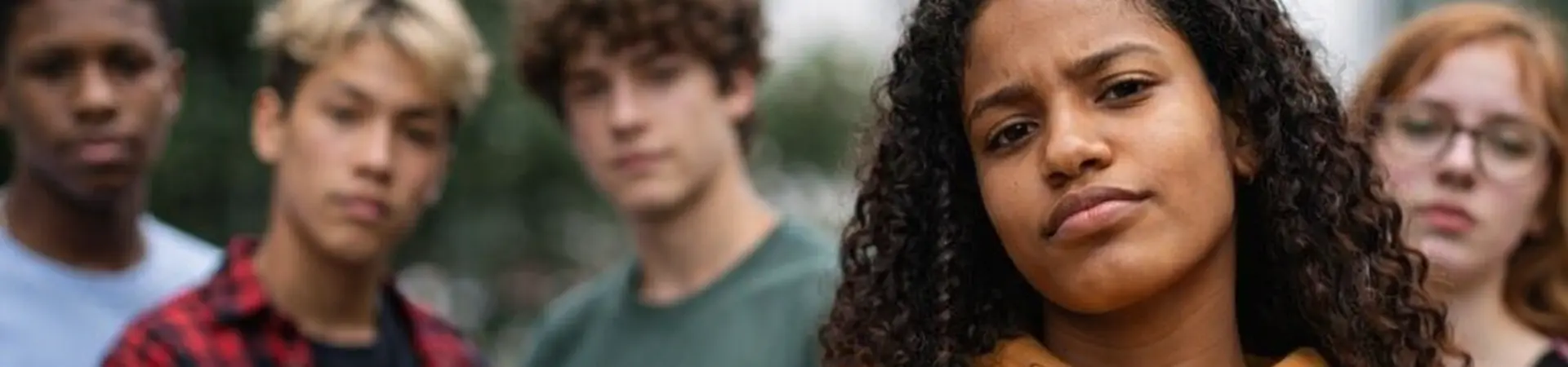 Five diverse young people stand outside, with a confident woman in a yellow hoodie at the forefront, all looking towards the camera.