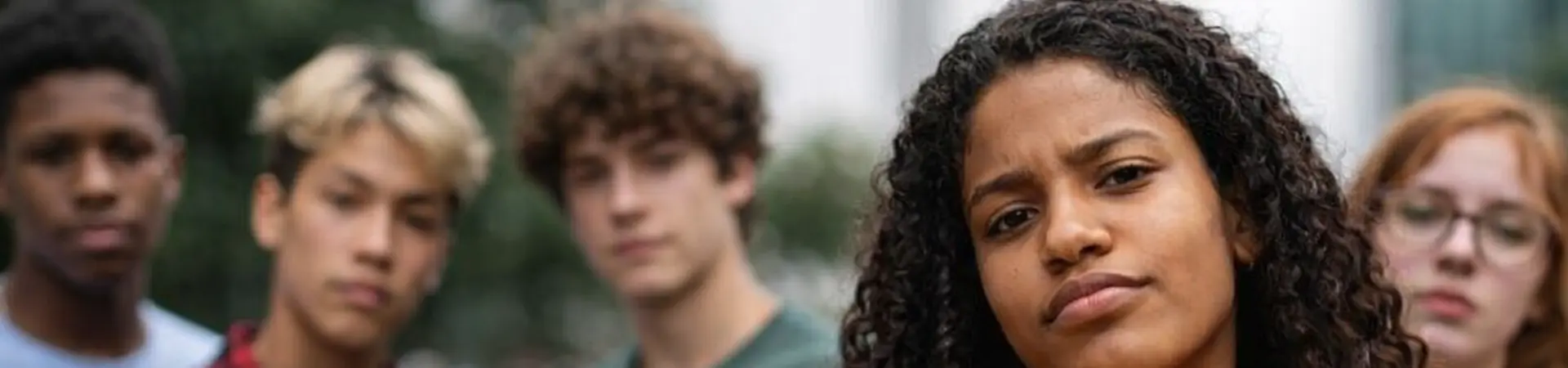 Five diverse young people stand outside, with a confident woman in a yellow hoodie at the forefront, all looking towards the camera.