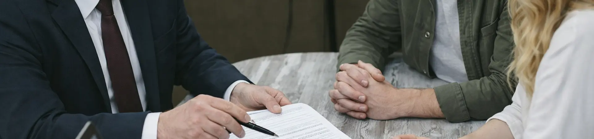 A businessman in a suit discusses a document with a man and woman at a table, with a laptop and plant nearby.