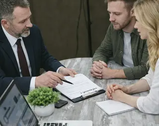 A businessman in a suit discusses a document with a man and woman at a table, with a laptop and plant nearby.