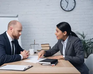 Two professionals in suits discussing paperwork at a table, with a flip chart, books, and a clock in the background.